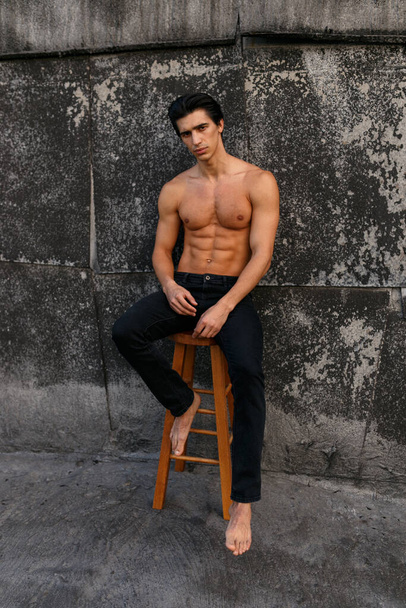Full length portrait of attractive young man with naked torso near old black wall, seated on high chair. Sportive healthy man. - Foto, afbeelding