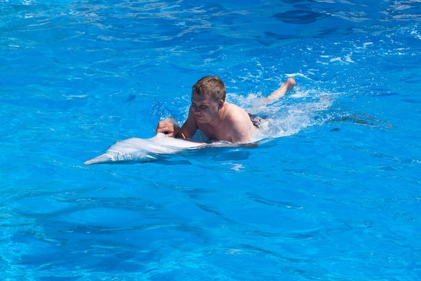 Egy fiatalember lovagol delfinen a hátán, fiú úszik delfinnel a kék vízben a medencében, a tengerben, az óceánban, a delfin megment egy embert. - Fotó, kép