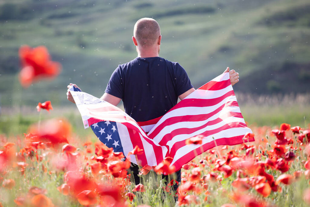 Attraktiver Mann mit der Flagge der Vereinigten Staaten in einem schönen Mohnfeld an einem klaren, sonnigen Tag. Feiern zum Unabhängigkeitstag, Konzept zum Nationalfeiertag. - Foto, Bild