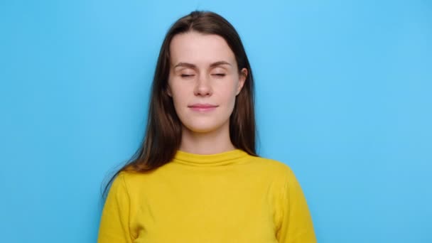 Portrait of glad beautiful young woman shows heart gesture over chest, being passionate, express love to close person, wears yellow jumper, stands over blue studio background. Sincere confession - Imágenes, Vídeo