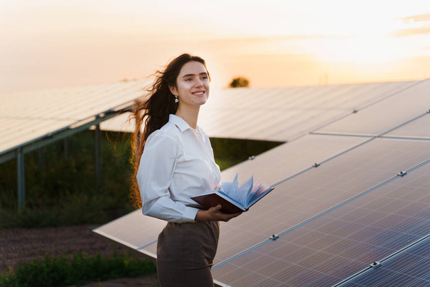 Girl and solar panels stands in row on the ground at sunset. Woman dressed white formal shirt on the power plant. Free electricity for home. Green energy. Solar cells power plant business. - Photo, Image