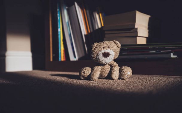 Teddy bear is sitting down on carpet next to blurry bookshelf background in retro filter,Lonely teddy bear stay home alone in living room at night, lonely concept, international missing children's day - Photo, Image