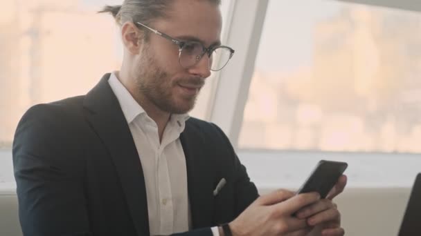 A good-looking young bearded businessman is using his smartphone while working in a city cafe indoors - Video