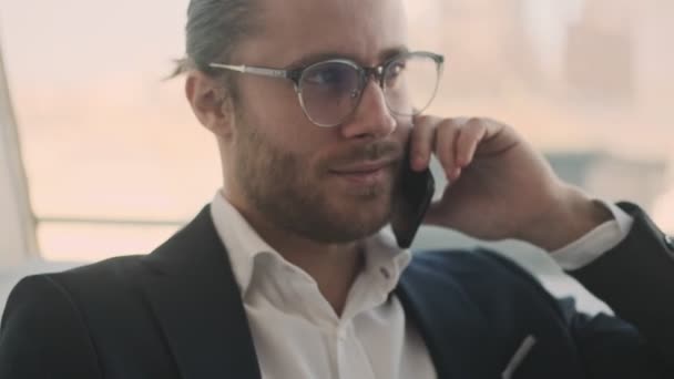 A handsome young bearded businessman is talking on his smartphone while working with paper documents in a city cafe indoors - Materiał filmowy, wideo