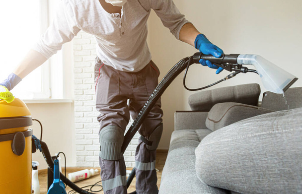 Cleaning service. Man janitor in gloves and uniform vacuum clean sofa with professional equipment - Photo, image
