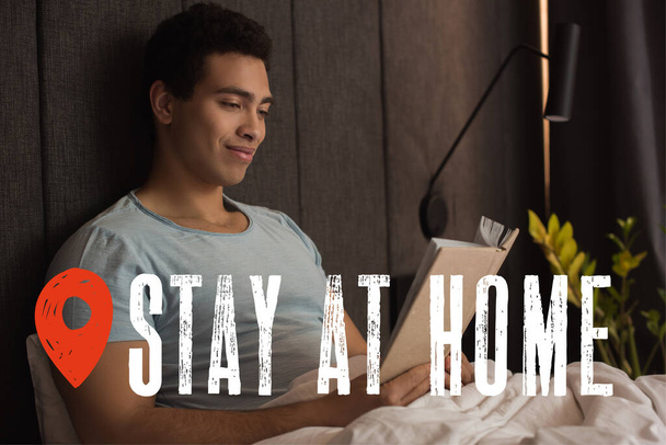 happy mixed race man reading book near stay at home lettering in bedroom  - Fotoğraf, Görsel