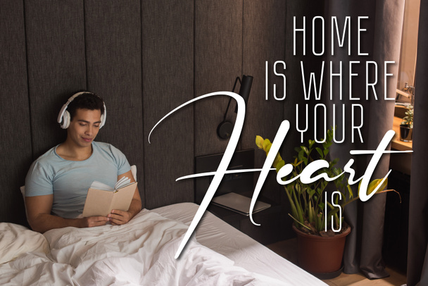 handsome mixed race man in headphones listening audio book and holding book near home is where your heart is lettering in bedroom  - Photo, Image
