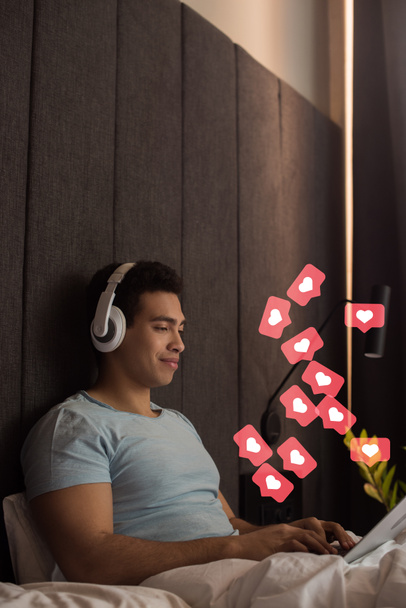 bi-φυλετικός άνθρωπος ακούγοντας μουσική στα ακουστικά και χρησιμοποιώντας φορητό υπολογιστή κοντά σε εικονικές καρδιές, όπως αρέσει στην κρεβατοκάμαρα  - Φωτογραφία, εικόνα