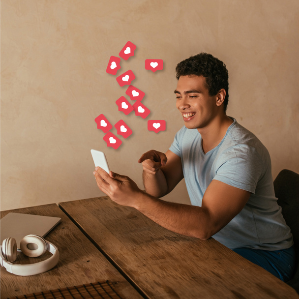 happy mixed race man pointing with finger at smartphone, laptop, headphones and virtual hearts illustration  - Photo, Image