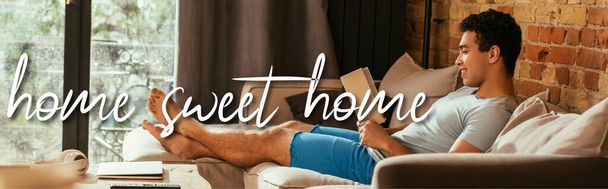 ホームスイートホームレタリングの近くのソファの上で本を読んでいる混合レースマンの笑顔のウェブサイトヘッダー  - 写真・画像