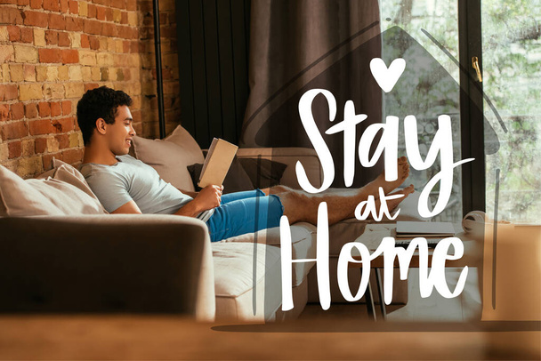 happy mixed race man reading book near stay at home lettering in living room  - Photo, Image
