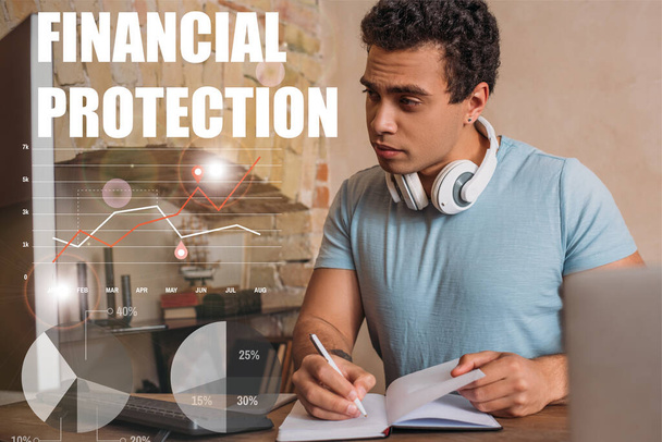 selective focus of handsome mixed race freelancer writing in notebook near computer monitor and financial protection lettering  - 写真・画像