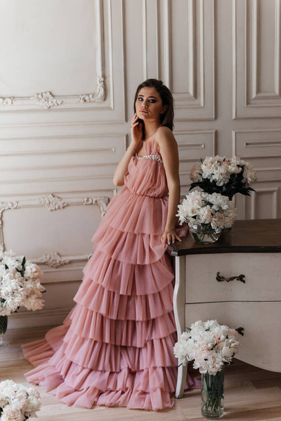 photo in the style of advertising expensive perfumes. a girl in a vintage studio dressed in a beautiful pink dress and surrounded by bouquets of fresh peonies - Photo, Image