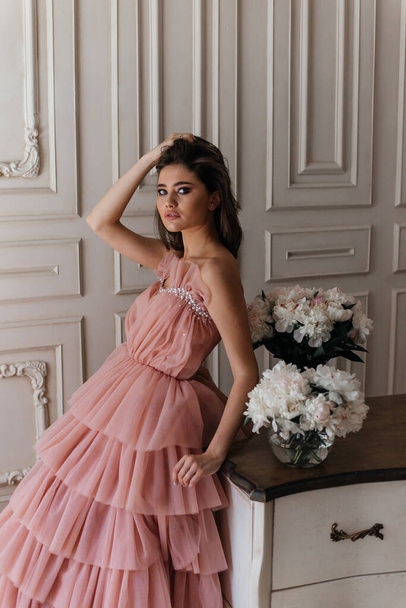 photo in the style of advertising expensive perfumes. a girl in a vintage studio dressed in a beautiful pink dress and surrounded by bouquets of fresh peonies - Фото, изображение