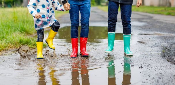 Üç çocuk, yeni yürümeye başlayan bir kız ve iki çocuk kırmızı, sarı ve yeşil çizme giyiyorlar ve yağmurluk sırasında yürüyorlar. Mutlu kardeşler gölete atlıyor. Dışarıda eğleniyor, aktif bir aile. - Fotoğraf, Görsel