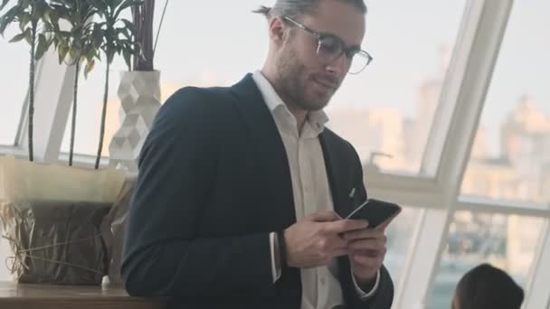 A pleased businessman is using his smartphone while standing and waiting in a city cafe indoors - Video