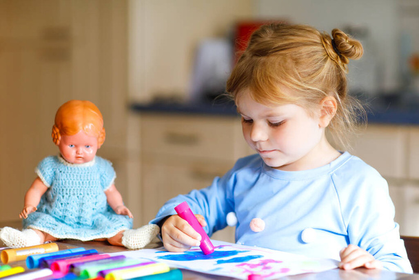 little alone toddler girl painting with felt pens during pandemic coronavirus quarantine disease. Happy creative child with old vintage doll, homeschooling and home daycare with parents - Photo, Image