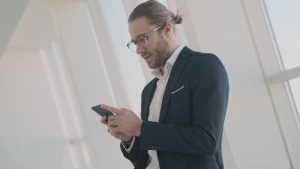 A happy smiling young businessman is using his smartphone while standing in a city cafe near windows indoors - Filmmaterial, Video