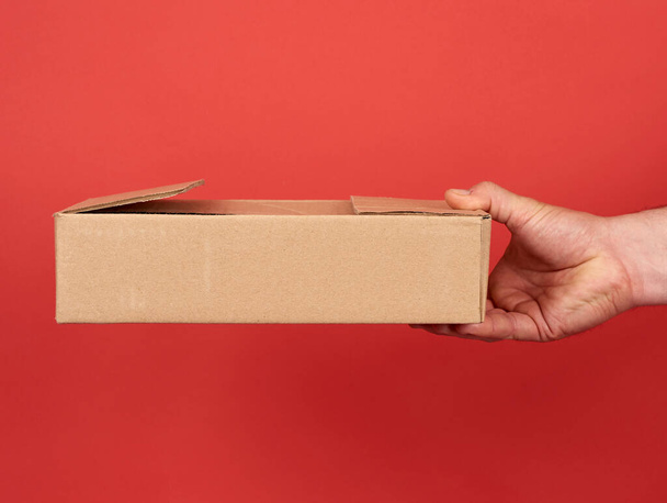 mannelijke hand houdt een bruine kartonnen doos op een rode achtergrond, concept van de levering van goederen - Foto, afbeelding