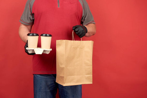 man in a red T-shirt, wearing black latex gloves, holds a tray with disposable cups of coffee and a paper bag from brown kraft paper, red background - Foto, Imagem