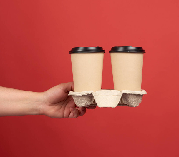 brown paper disposable cups with a plastic lid stand in the tray, female hand holds disposable tableware on a red background - Foto, Imagem