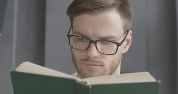 Close-up face of smart young man in eyeglasses reading book. Portrait of bearded brunette Caucasian reader enjoying literature indoors. Lifestyle, hobby, intelligence, education. Cinema 4k ProRes HQ. - Materiał filmowy, wideo