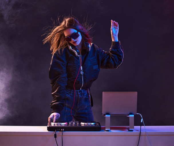 Stylish girl DJ in glasses headphones and a bomber mixes music dancing at a party in the neon light in the smoke . - Фото, зображення