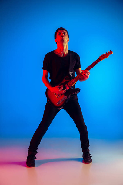 Young caucasian musician playing guitar in neon light on blue background, inspired - Foto, Bild