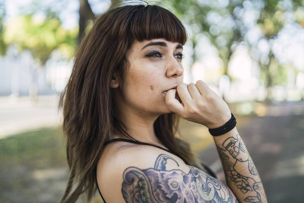 Una joven atractiva mujer caucásica con tatuajes de pie en el parque y haciendo una cara linda - Foto, imagen