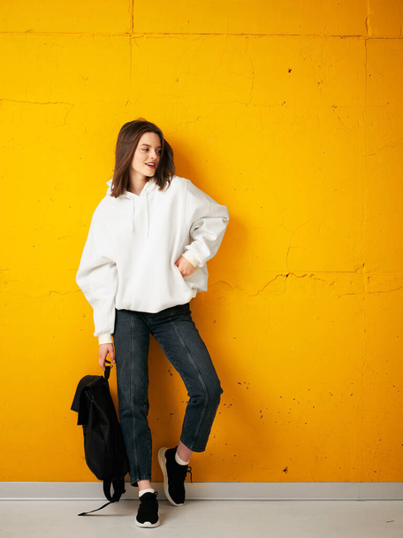 Portrait of a young pretty girl student in a white hoodie, casual clothes, with a bag, against a concrete orange wall. - Foto, Imagen