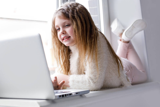 The girl at home communicates with friends on the Internet. Kids distance learning. Cute little girl using laptop at home. Education, online study, home studying, schoolgirl children lifestyle concept - Foto, afbeelding