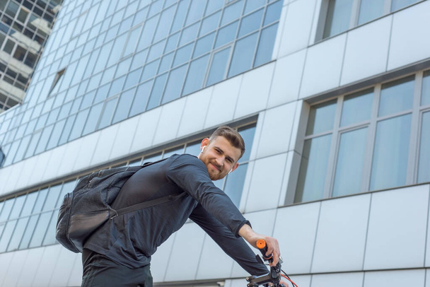 オフィスビルの高層ビルを背景にカメラを見て笑っている若い男が自転車に乗って自転車に乗りました。屋外だ。春のスポーツ。その男は２５歳です。サイクリング春の自転車に乗る。極端なサイクリング。アクティブなライフスタイル - 写真・画像