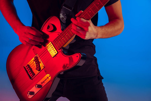 Young caucasian musician playing guitar in neon light on blue background, inspired - Foto, Bild