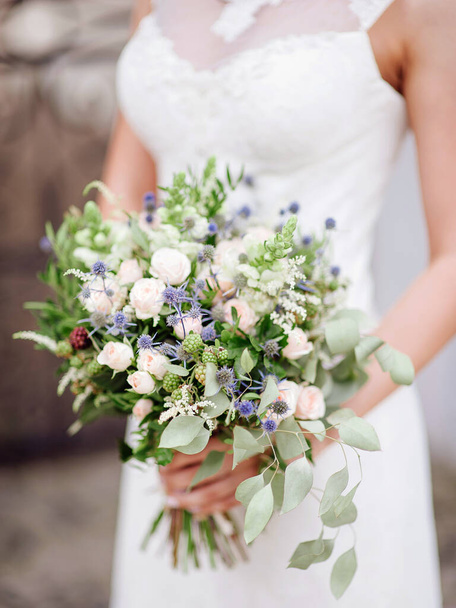 Νεαρή νύφη κατέχει υπερμεγέθης γαμήλια ανθοδέσμη με μπλε γαϊδουράγκαθο λουλούδια, jana τριαντάφυλλα, φύλλα ευκαλύπτου κοντά. Θερινή γαμήλια τελετή.  - Φωτογραφία, εικόνα