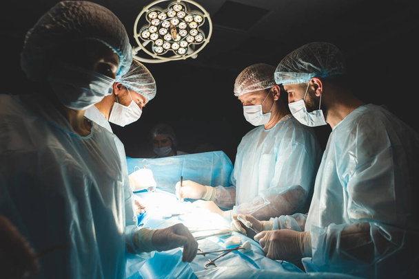An international professional team of surgeon, assistants and anesthesiologist perform a complex operation on a patient under general anesthesia. Dark atmospheric photography theme in low key. - Photo, image