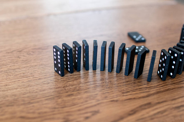 schwarze Dominokarten mit weißen Punkten in Reihen, die nacheinander auf einem Holztisch balanciert werden. Fliesen aus dem Dominostein, bevor sie einem Ketteneffekt zum Opfer fallen. Geschäftskatastrophen-Konzept mit Dominoeffekt - Foto, Bild