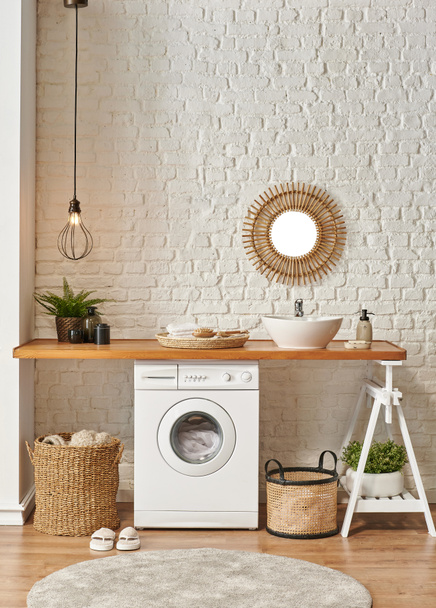 Máquina de lavar roupa na lavanderia, mesa de madeira e estilo de prateleira, espelho de lâmpada de pia e objeto de decoração de cesta de vime
. - Foto, Imagem
