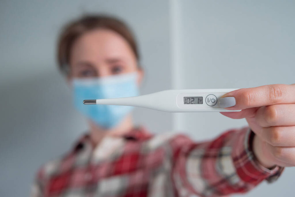 Mujer en mascarilla que muestra termómetro médico con alta temperatura corporal - Foto, imagen
