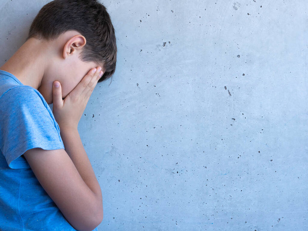 Αναστατωμένο θλιμμένο αγόρι στέκεται μόνο του και ακουμπά στον γκρίζο τοίχο. Μαθησιακές δυσκολίες, οικογενειακά προβλήματα, εκφοβισμός, κατάθλιψη, άγχος ή απογοήτευση - Φωτογραφία, εικόνα
