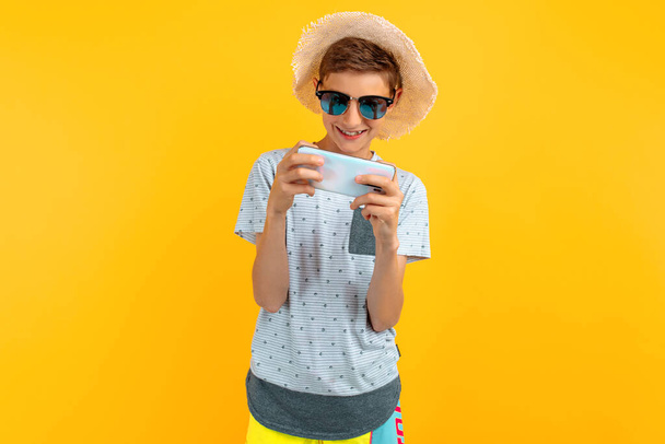 Stylish smiling teen guy in a summer hat using a mobile phone standing on an isolated yellow background - Fotó, kép
