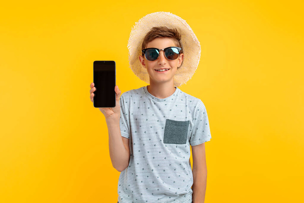 Happy smiling teen guy in a summer hat, shows a blank mobile phone screen for a copy of the space, on an isolated yellow background - Fotó, kép