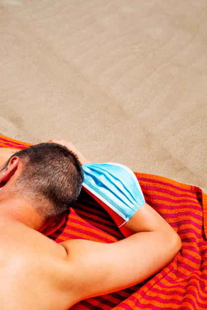 close-up de um homem caucasiano, deitado de cara para baixo em uma toalha de laranja colorida na praia, vestindo uma máscara cirúrgica azul em seu braço, como está tomando um fôlego de usá-lo
 - Foto, Imagem