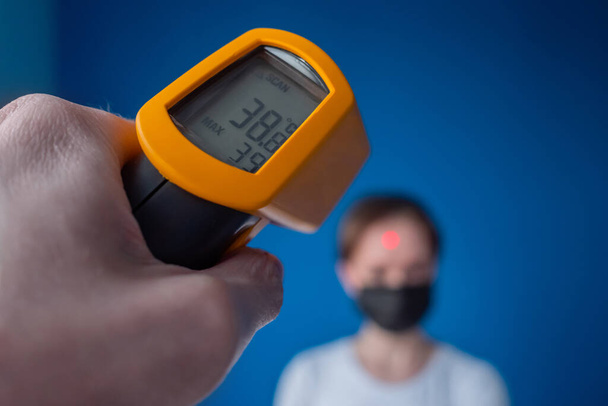 Man hand holding pyrometer, measuring temperature of woman in medical face mask - Photo, Image