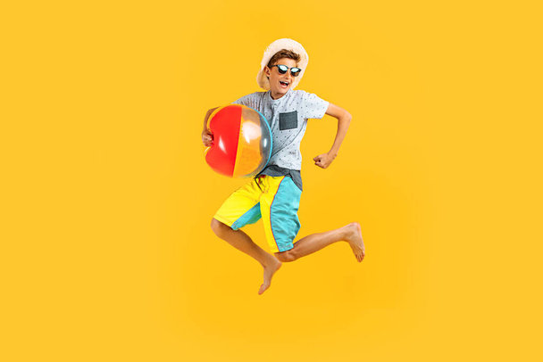 Full-length portrait of a happy excited teenager in sunglasses and a summer hat, having fun and jumping, holding an inflatable sea ball on a yellow background - Foto, imagen