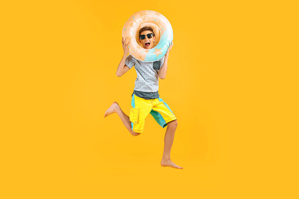 Full-length portrait of a happy excited teenager in sunglasses and a summer hat, having fun and jumping, holding an inflatable ring on a yellow background - 写真・画像