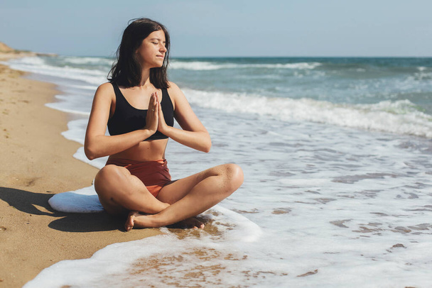 Joven hermosa mujer practicando yoga en la playa, sentada en la arena y meditando. Salud mental y autocuidado. Chica feliz relajarse en la orilla del mar en las vacaciones de verano
 - Foto, Imagen