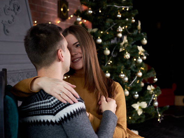 Young couple in love sitting by fireplace near Christmas tree, kissing on a date. Handsome brunette man, wearing grey winter sweater kissing pretty woman in mustard yellow knitted dress. - Fotoğraf, Görsel