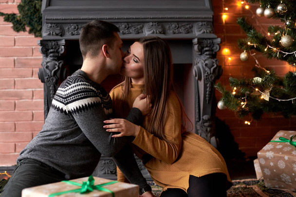 Young couple in love sitting by fireplace near Christmas tree on a date with presents. Handsome brunette man, wearing grey winter sweater kissing pretty woman in mustard yellow knitted dress. - Photo, image