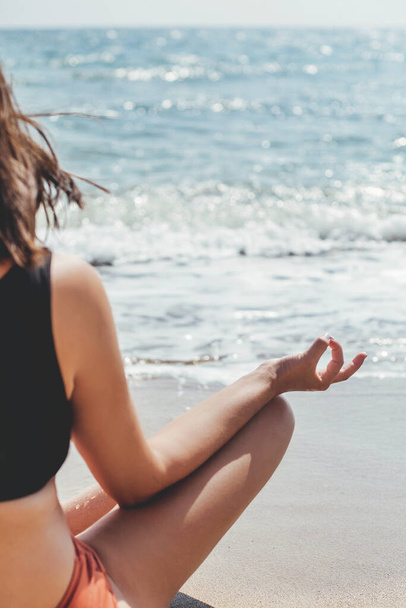 Young beautiful woman practicing yoga on the beach, sitting on sand and meditaning at sea. Mental health and self care. Happy girl relaxing on seashore on summer vacation, cropped view - Photo, Image