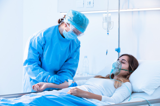 Νοσοκόμα που ελέγχει μια ενδοφλέβια έγχυση σε ασθενή που φοράει μάσκα οξυγόνου θετικής πίεσης στο νοσοκομείο κατά τη διάρκεια της πανδημίας του Covid- 19 - Φωτογραφία, εικόνα